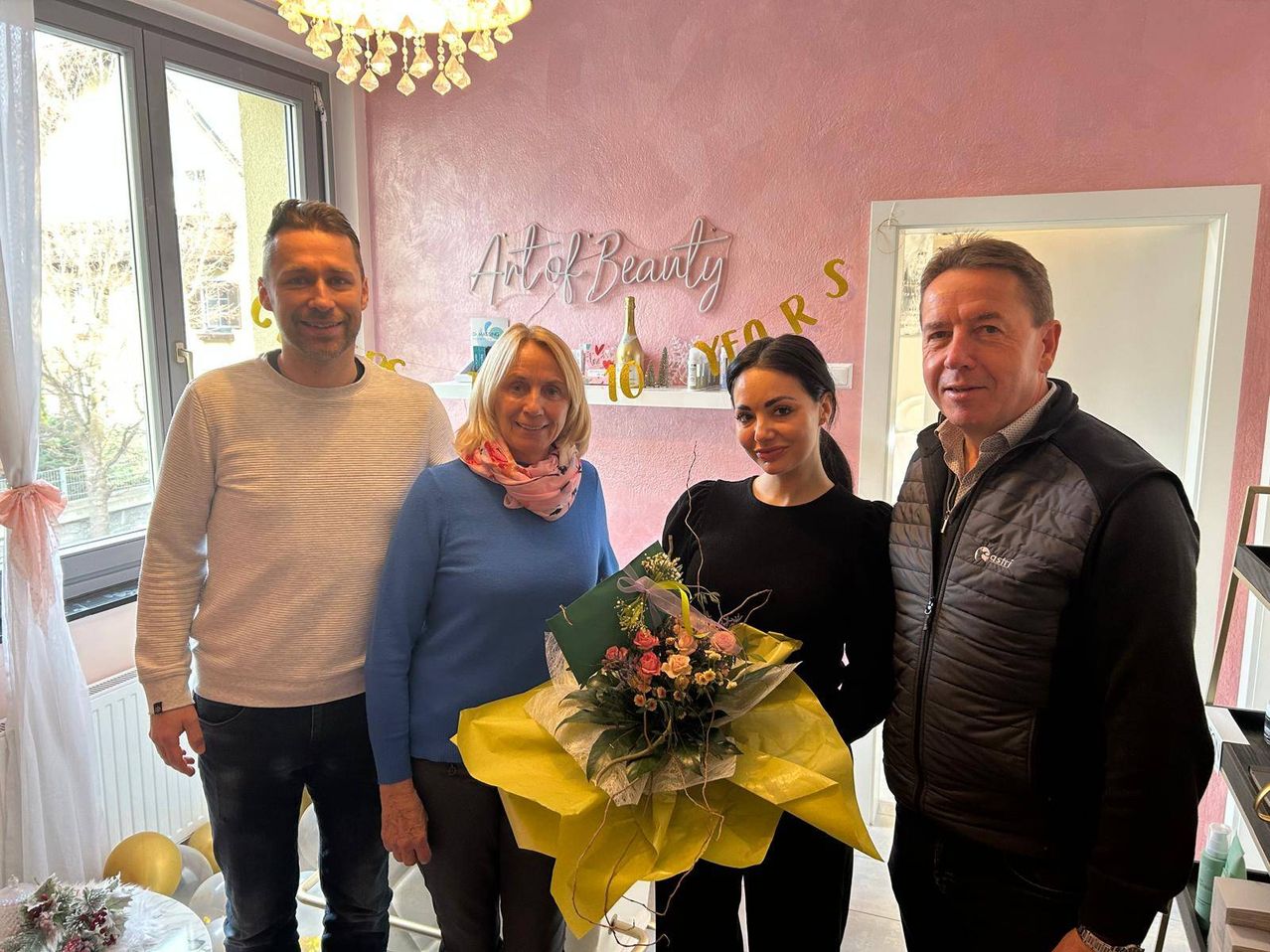 Vier Personen stehen in einem Schönheitssalon und lächeln für ein Foto. Eine Frau hält einen Blumenstrauß. An der rosa Wand hinter ihnen steht 'Art of Beauty' und '10 JAHRE' geschrieben.