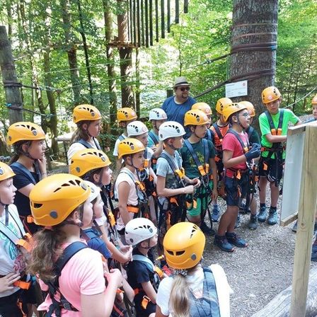 Eine Gruppe von Kindern und Erwachsenen mit gelben Helmen und Gurten versammelt sich in einem Wald, wahrscheinlich in Vorbereitung auf eine Abenteueraktivität.