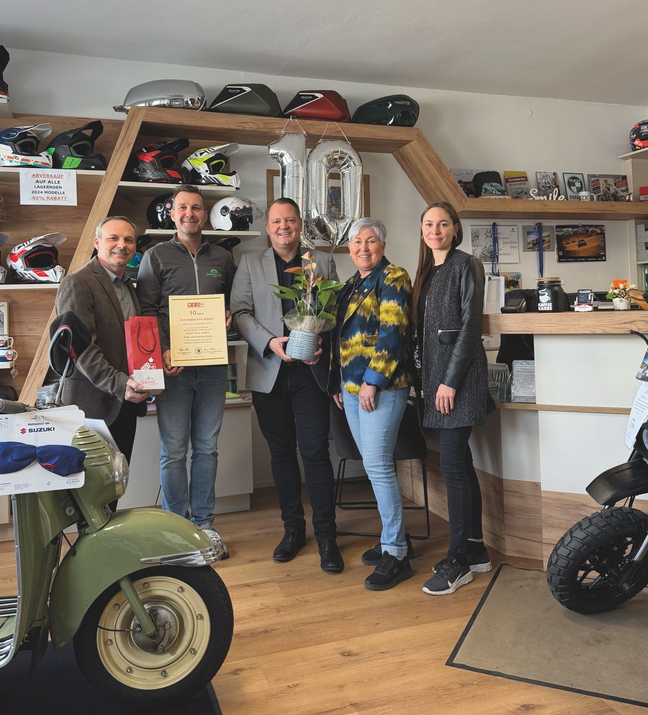 Fünf Personen stehen in einem Geschäft mit ausgestellten Rollern und Helmen. Ein Mann hält ein Zertifikat und eine Frau eine Topfpflanze. Der Laden hat Holzregale, eine weiße Theke und Luftballons.