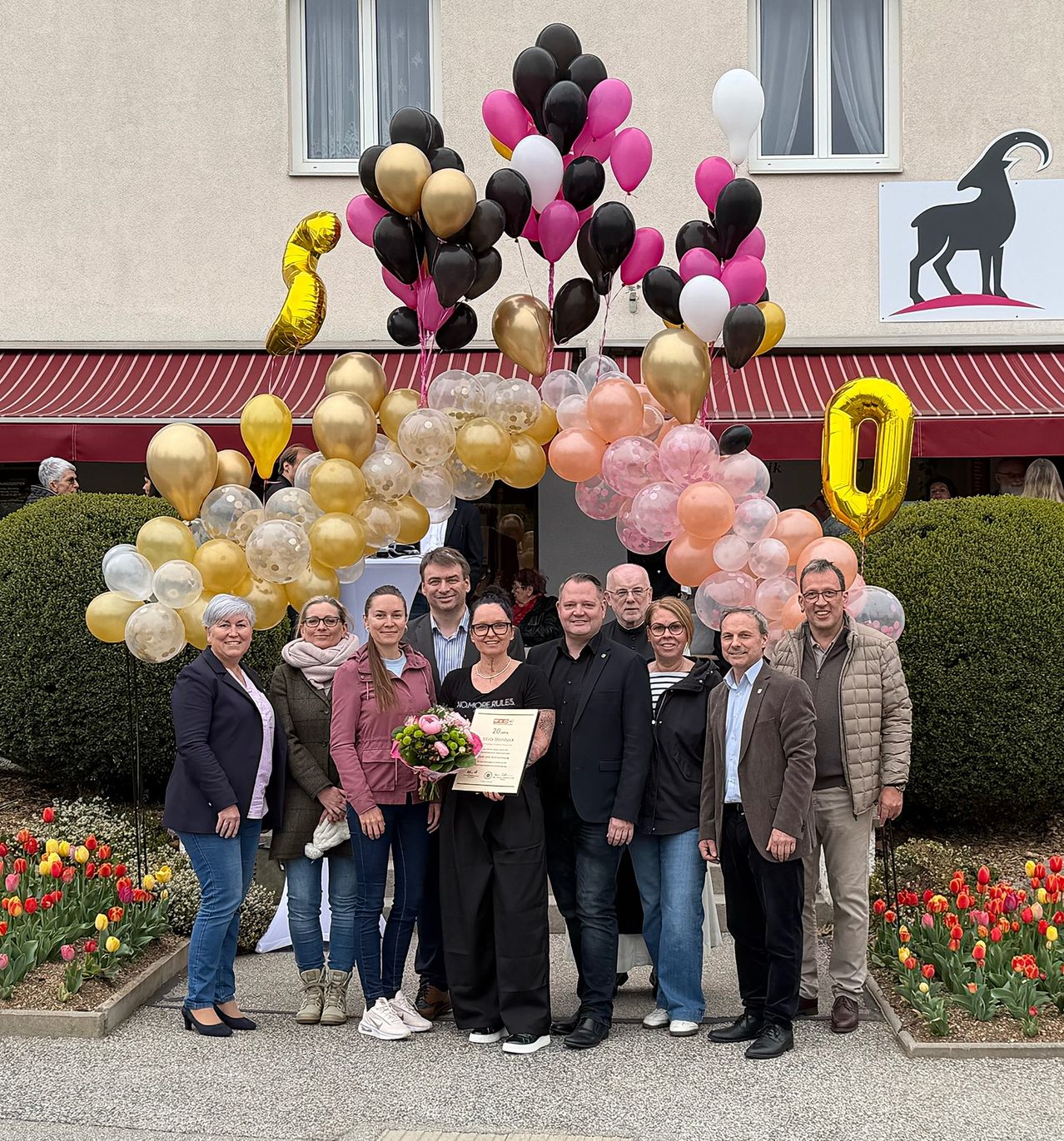 Eine Gruppe von Menschen posiert für ein Foto vor einem Gebäude mit Luftballons. Sie lächeln und halten ein Zertifikat. Das Gebäude hat ein Schild und ein Fenster. Es gibt Blumen auf dem Boden.