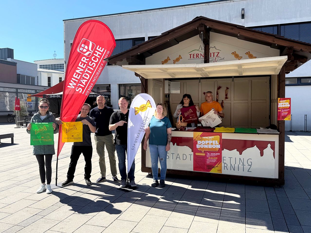 Fünf Personen stehen vor einem Kiosk und halten Banner. Eine Frau in der Mitte hält ein rotes Schild, während zwei Frauen im Kiosk einen Korb und ein Schild halten. Der Kiosk hat ein Schild mit der Aufschrift Doppel Bonbon.