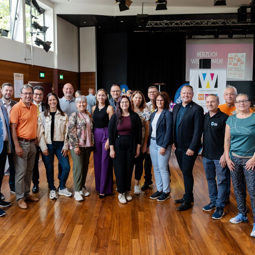 Eine Gruppe von Menschen, sowohl Männer als auch Frauen, stehen zusammen auf einem Holzboden, wahrscheinlich für ein Gruppenfoto. Sie alle lächeln und scheinen für die Kamera zu posieren. Hinter ihnen befindet sich eine Bühne mit einer Projektionsleinwand und einem Banner.