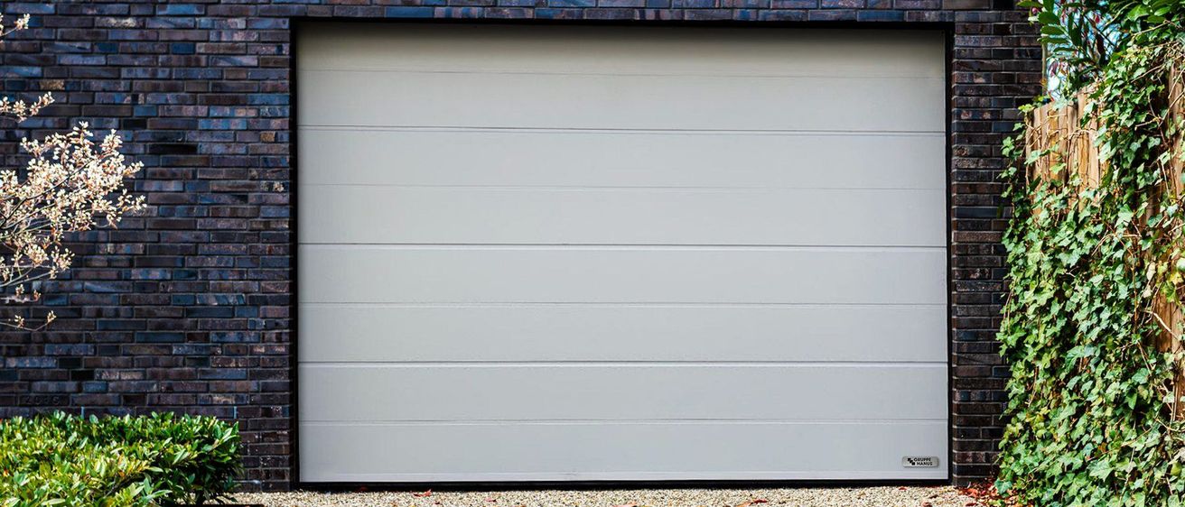 Eine weiße Garagentür mit einem silbernen Griff ist an einer Ziegelwand montiert, mit Bäumen im Hintergrund.