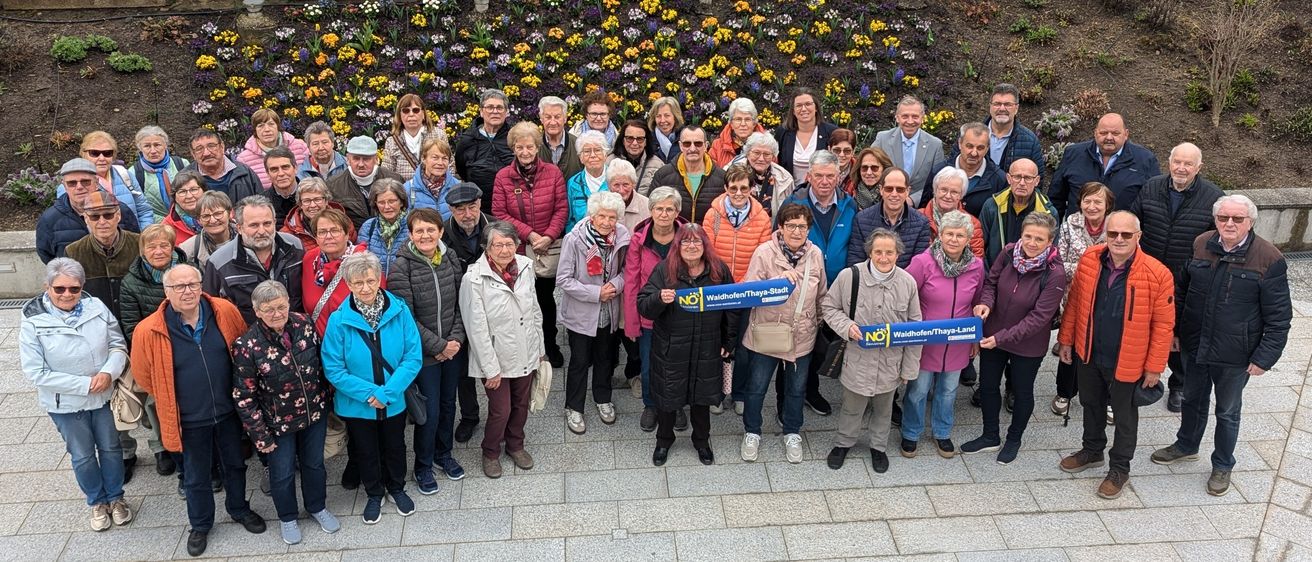 Eine Gruppe älterer Personen, alle in Winterkleidung, posiert für ein Foto vor einer mit Blumen geschmückten Wand. Einige von ihnen halten Schilder mit der Aufschrift 'NEIN'.