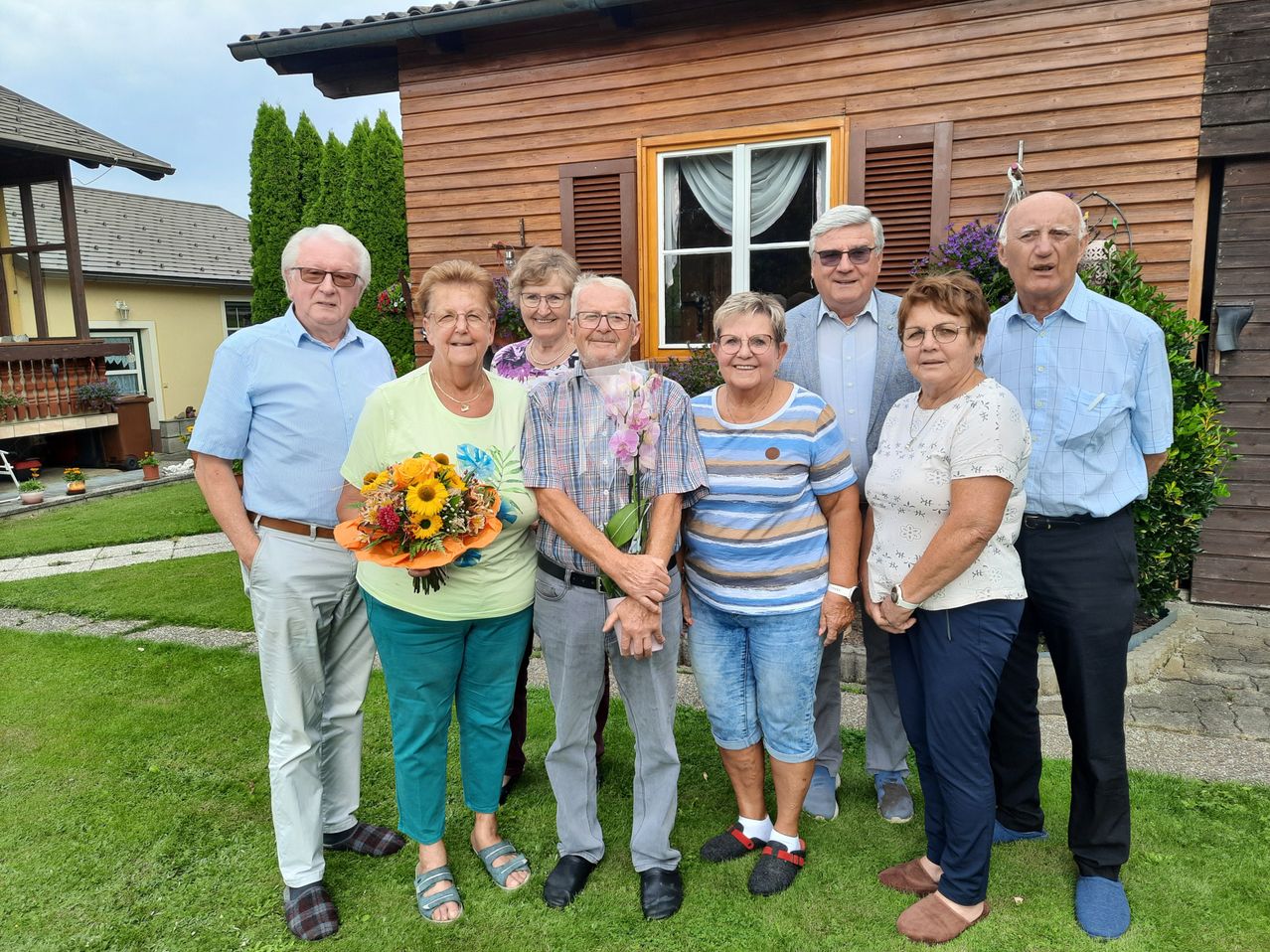 Eine Gruppe älterer Erwachsener steht auf dem Rasen vor einem Holzhaus. Eine Frau hält einen Blumenstrauß.