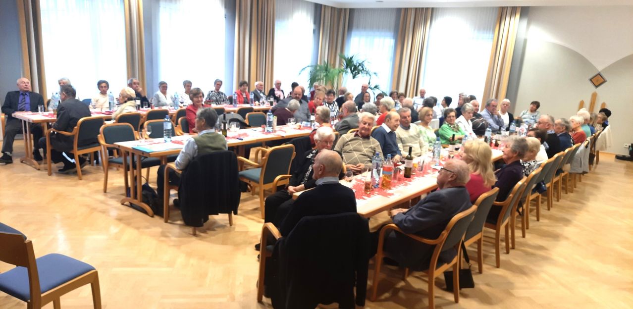 Eine Gruppe älterer Menschen sitzt an Tischen in einem Konferenzraum, mit Weinflaschen und Gläsern auf den Tischen und einer Pflanze in der Ecke.
