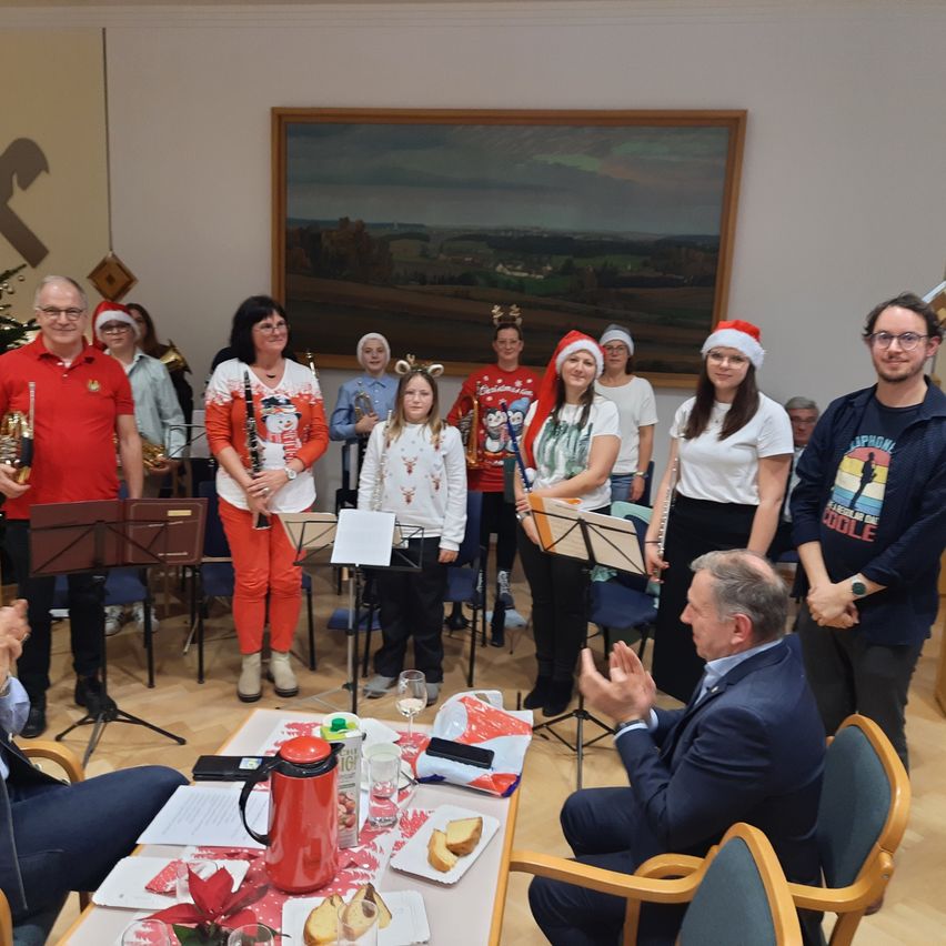 Eine Gruppe von Menschen in Weihnachtskleidung steht um einen Tisch, einige halten Musikinstrumente. Ein Mann klatscht, und andere hören zu. Der Hintergrund zeigt ein Gemälde an der Wand.