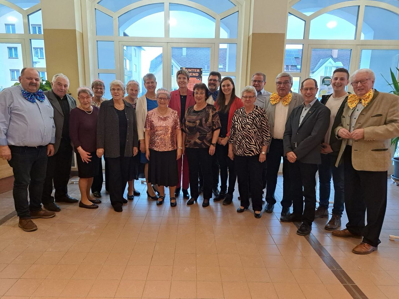 A group of people, mostly older adults, stands together in a room with large windows. They appear to be posing for a photo.
