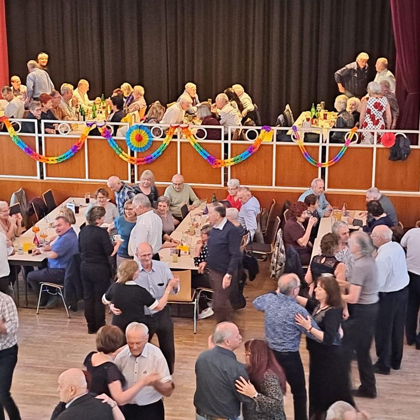 A gathering of older people inside a hall. They are sitting and standing around tables, possibly engaged in a social event. The room has wooden floors and a stage with a curtain. Colorful decorations adorn the area.