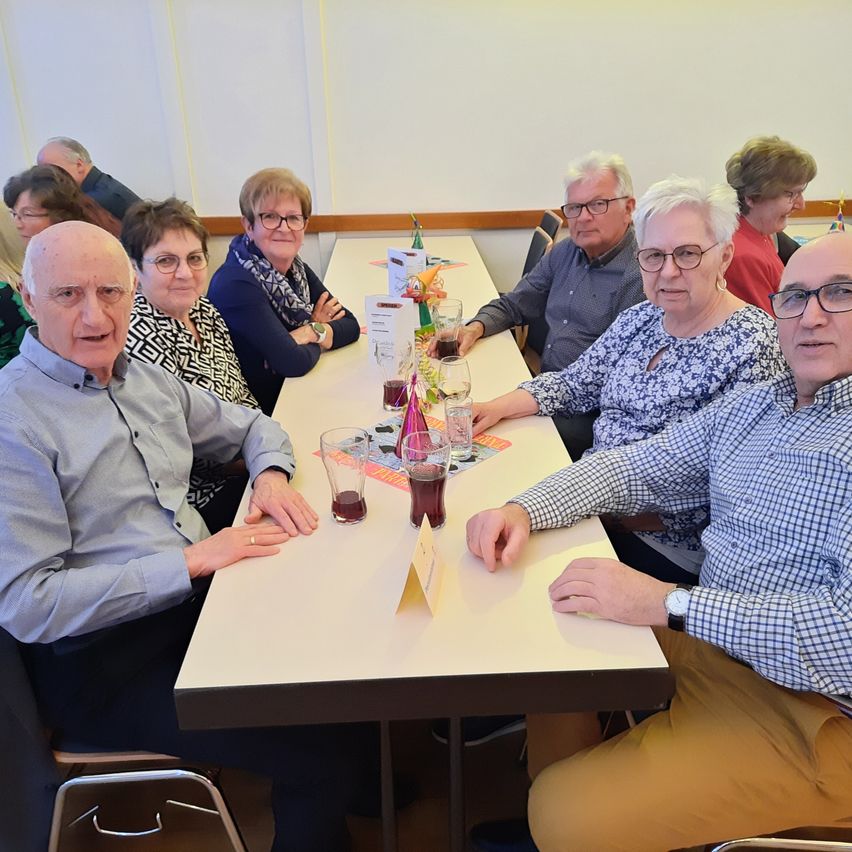 A group of elderly people are seated around a table in a room, possibly at a gathering. They are wearing glasses and some are wearing scarves. The table is set with cups, glasses, and plates. The room has white walls and a wooden floor.