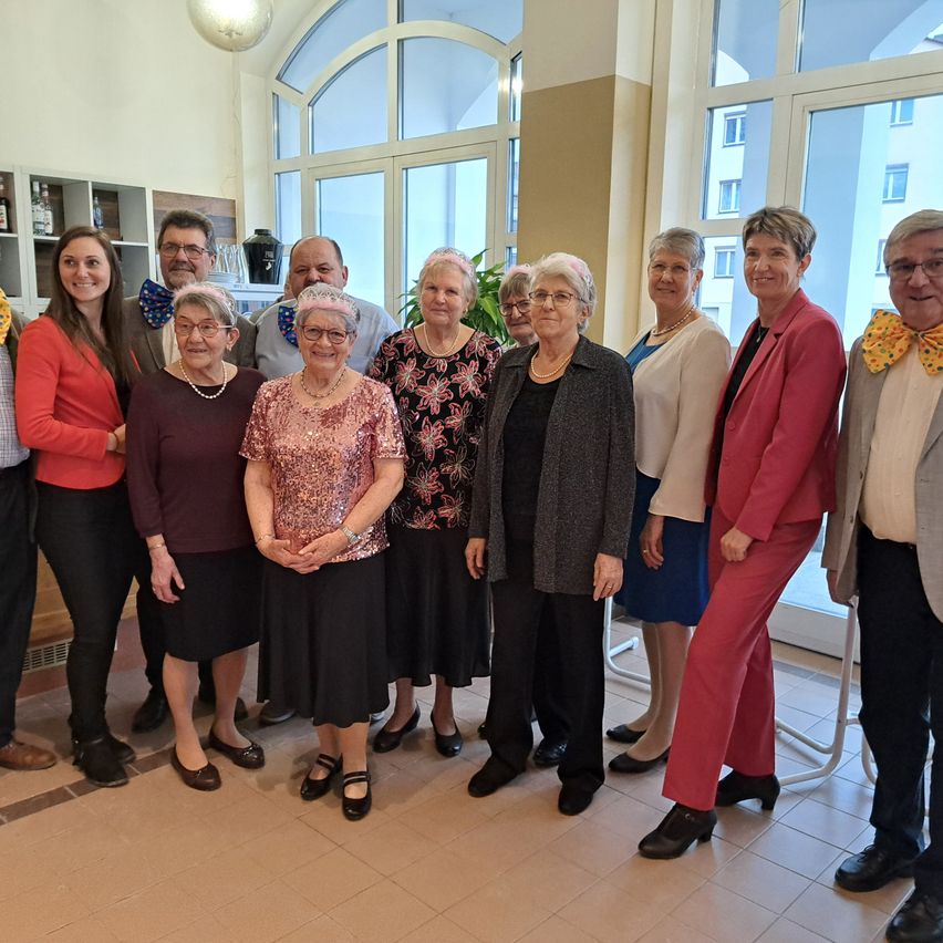 A group of people are standing together in a room. They are all wearing formal attire. Some of them are wearing glasses and necklaces. Behind them is a glass window that overlooks a building.