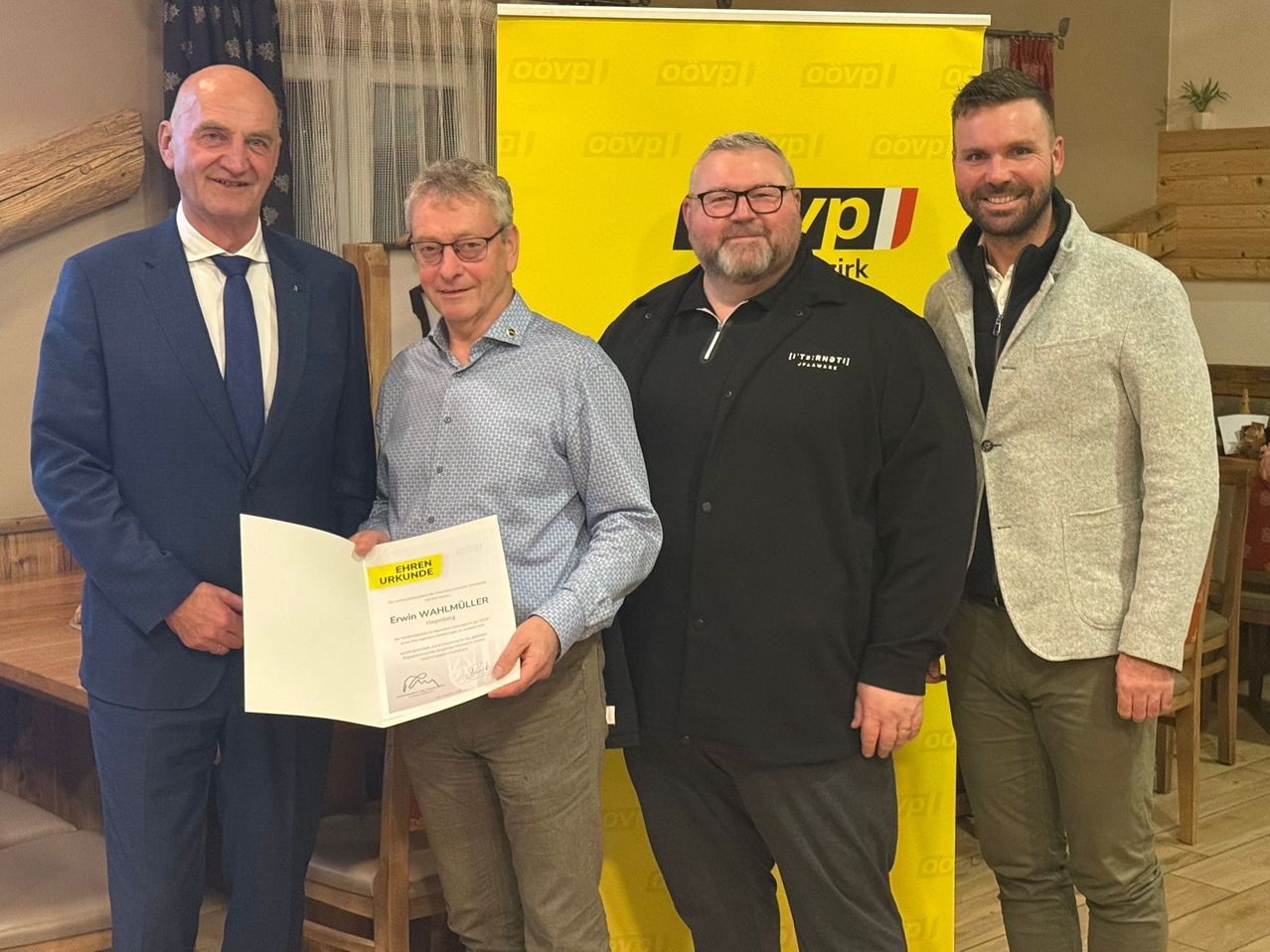 Four men stand in front of a yellow banner, one holding a certificate, while two others wear glasses and one has a beard. They are all smiling for the camera.