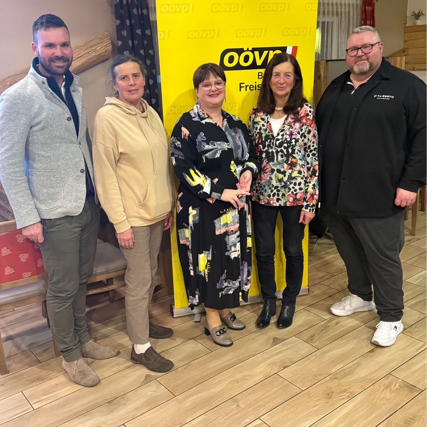 Five adults pose for a photo in front of a yellow banner with the text 'oov'. The woman in the middle wears glasses. Behind them is a wooden wall with a red chair.