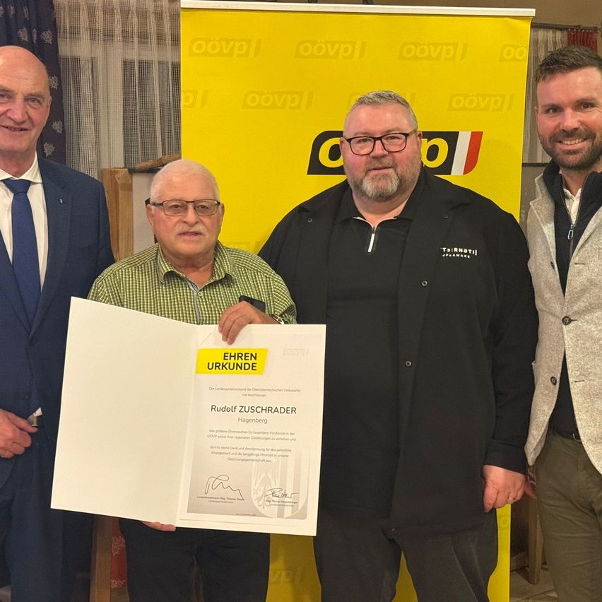 Four men stand in front of a yellow banner. The man in the center holds a certificate, and the man on the left smiles. Behind them is a wooden structure.