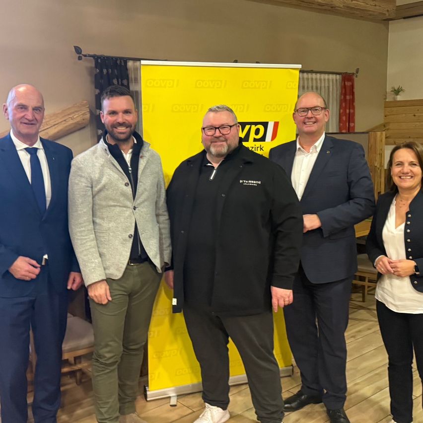 Five people are posing for a photo. They are standing in front of a yellow banner. The man in the middle is wearing glasses and a black jacket. The man on his right is wearing a suit and leather shoes. The man on his left is wearing a suit and a tie.