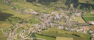 Luftaufnahme einer Stadt umgeben von grünen Feldern und bewaldeten Hügeln. Die Stadt hat ein Raster-Straßennetz mit vielen Gebäuden und Straßen. Es gibt ein großes Sportfeld und eine Autobahn, die durch die Landschaft führt.