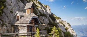 Eine kleine Holzkapelle auf einem steilen Bergfelsen, mit einem hölzernen Steg, der dorthin führt. Die Kapelle ist von üppigem Grün umgeben und bietet einen atemberaubenden Blick auf das Tal unten.