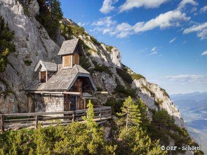 Eine kleine Holzkapelle auf einem steilen Bergfelsen, mit einem hölzernen Steg, der dorthin führt. Die Kapelle ist von üppigem Grün umgeben und bietet einen atemberaubenden Blick auf das Tal unten.