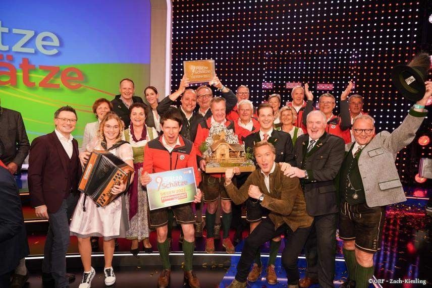 Eine Gruppe von Menschen steht auf einer Bühne und posiert für ein Foto mit Trophäen. Sie tragen traditionelle Kleidung und halten Schilder. Ein Mann spielt Akkordeon, und die Bühne hat einen farbenfrohen Hintergrund.