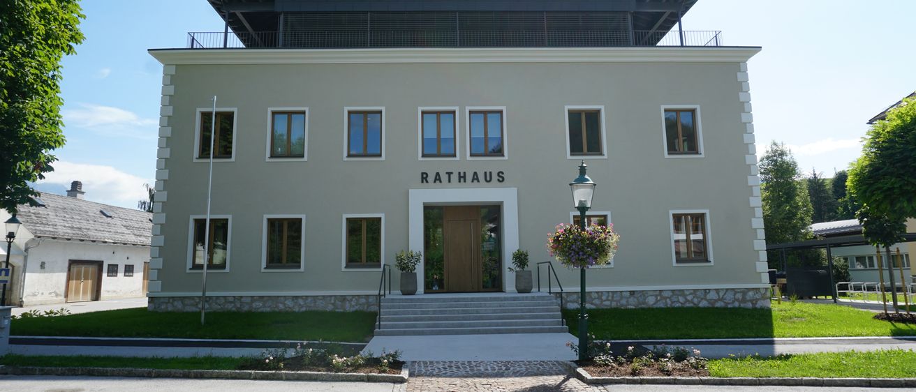Das Rathaus-Gebäude, ein beiges Gebäude mit schwarzem Dach, verfügt über einen Balkon, mehrere Fenster und einen zentralen Eingang mit Stufen und Topfpflanzen.