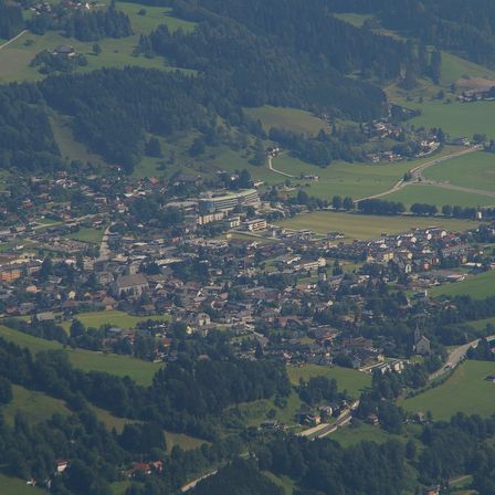 Luftaufnahme einer kleinen Stadt in einem Tal, umgeben von üppigen grünen Feldern und dichten Wäldern. Gebäude verschiedener Größen und Stile sind über das Gebiet verstreut.