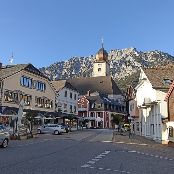 Eine Stadt mit Häusern und einer Kirche, Berge im Hintergrund. Mehrere Autos sind auf der Straße geparkt.