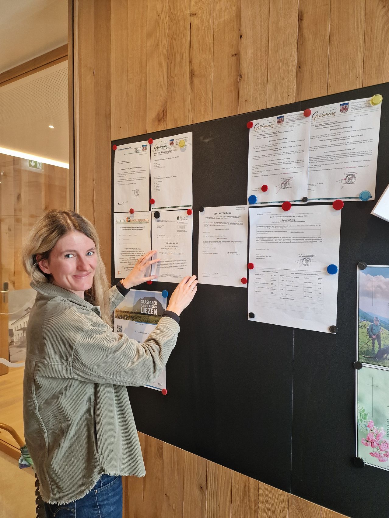 Eine Frau steht vor einer Pinnwand mit verschiedenen Dokumenten, lächelt und zeigt auf eines. Die Tafel ist an einer Holzwand mit einem Glasfenster in der Nähe befestigt.