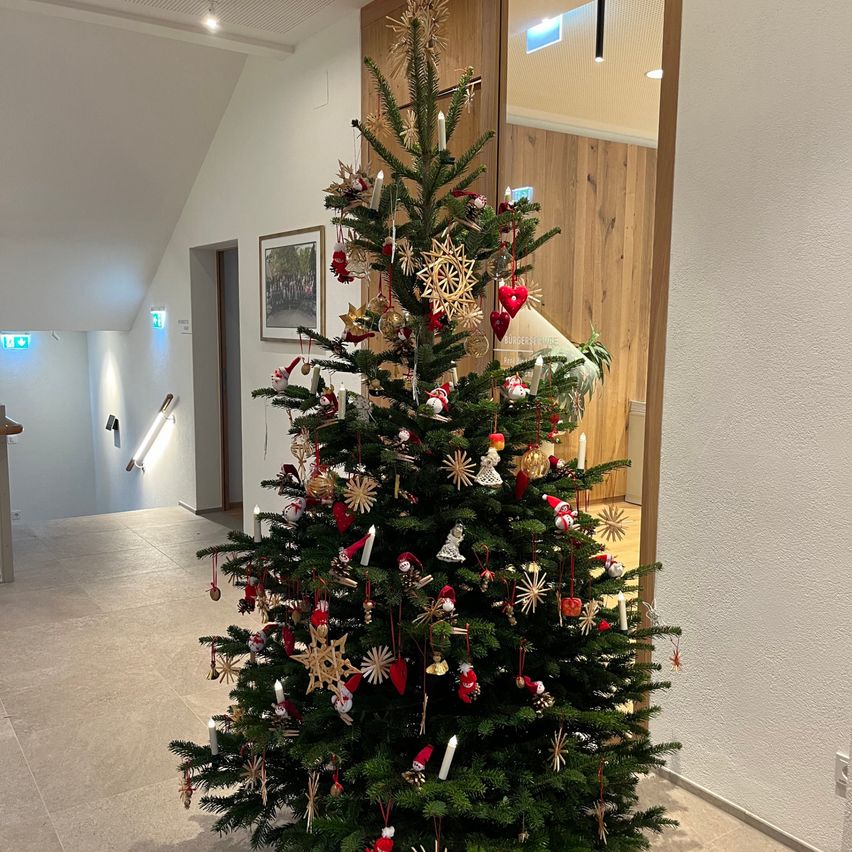 Ein Weihnachtsbaum steht in einem Flur, geschmückt mit Ornamenten und Kerzen. Dahinter spiegelt sich eine Holzwand in einer Glastür. Der Flur hat eine weiße Decke und einen gefliesten Boden.