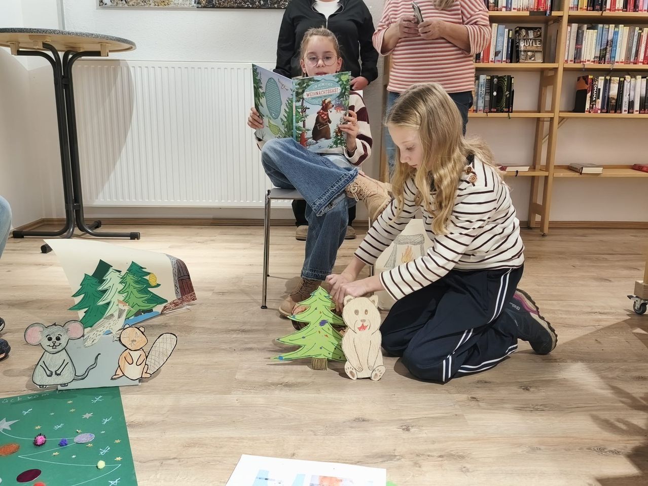 Drei Mädchen sind drinnen. Eine sitzt auf einem Stuhl und liest ein Buch, eine andere ordnet Spielzeug an und die dritte steht neben einem Bücherregal.