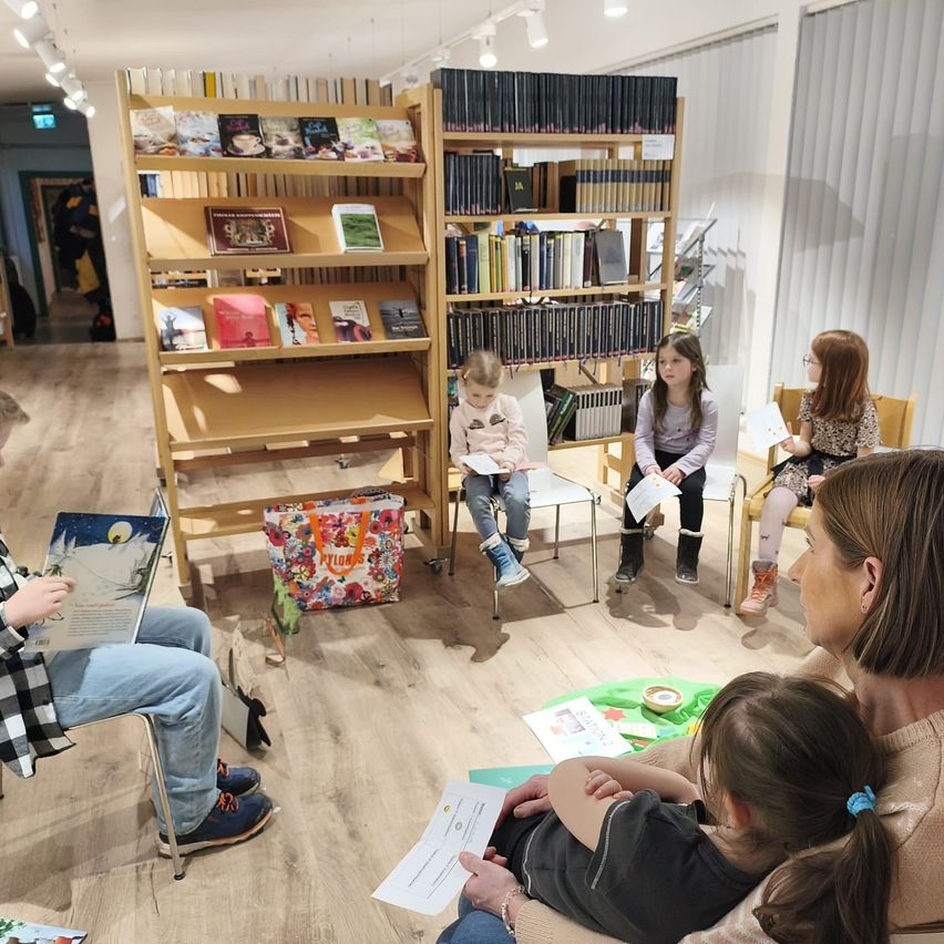 Eine Gruppe von Kindern und Erwachsenen sitzt in einem Kreis in einem Raum. Sie lesen Bücher. Eine Frau hält ein kleines Kind auf ihrem Schoß. Hinter ihnen stehen Regale, die mit Büchern gefüllt sind.