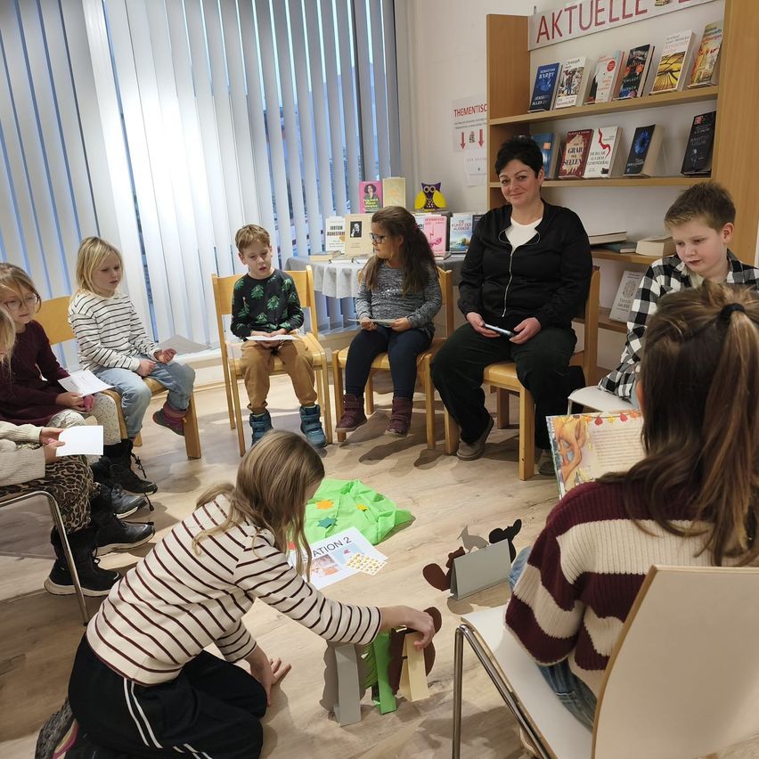 Eine Gruppe von Kindern sitzt in Stühlen um eine Frau in einer Bibliothek. Einige lesen Bücher und andere spielen mit Spielzeug. Der Raum hat Holzböden und Regale voller Bücher.