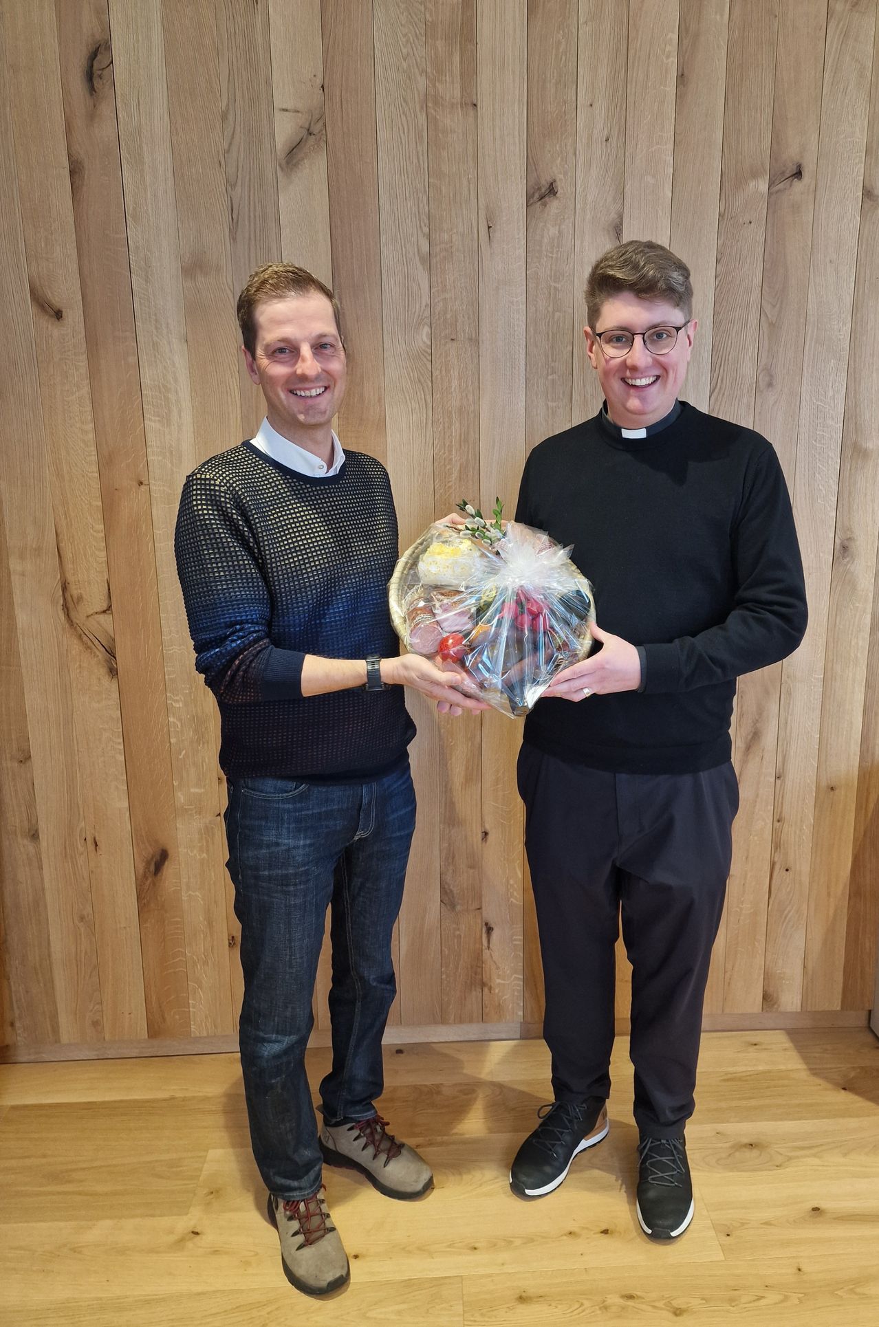 Zwei Männer stehen lächelnd da, einer hält einen verpackten Korb mit Obst und Gemüse vor einer Holzwand.