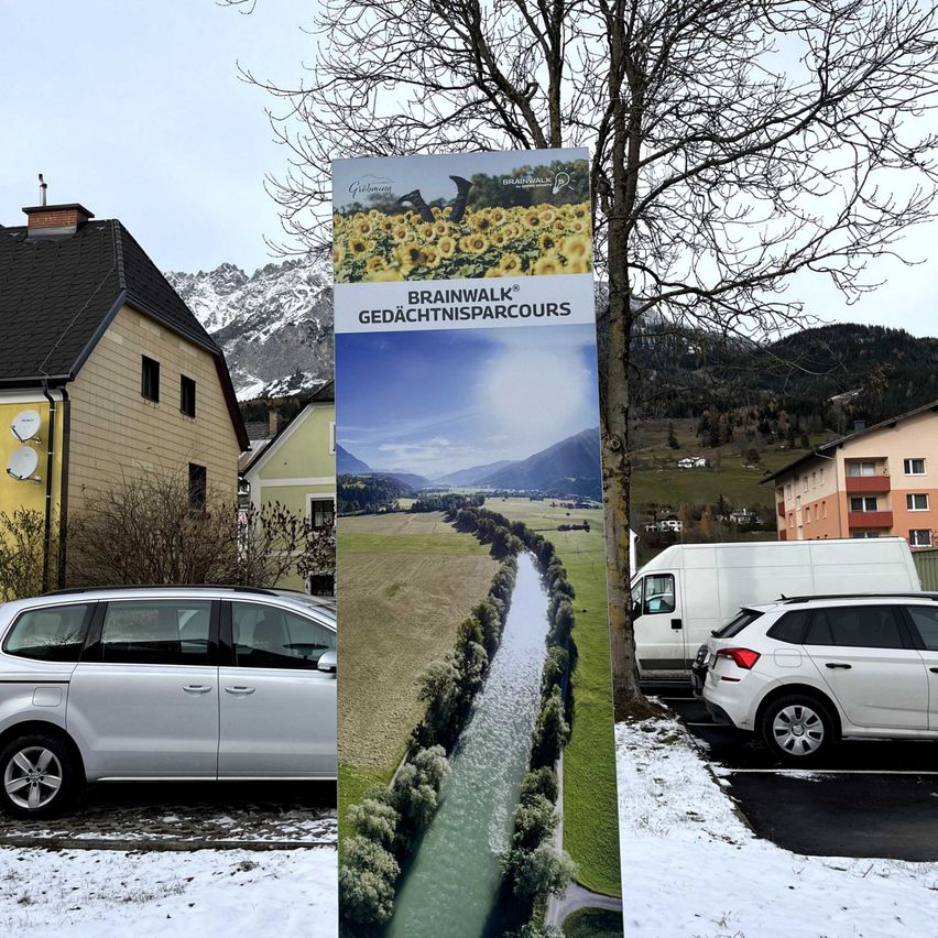 Ein Schild für einen BrainWalk-Gedachtnispfad steht in einer verschneiten Landschaft mit Häusern und Bergen. Autos sind in der Nähe geparkt.