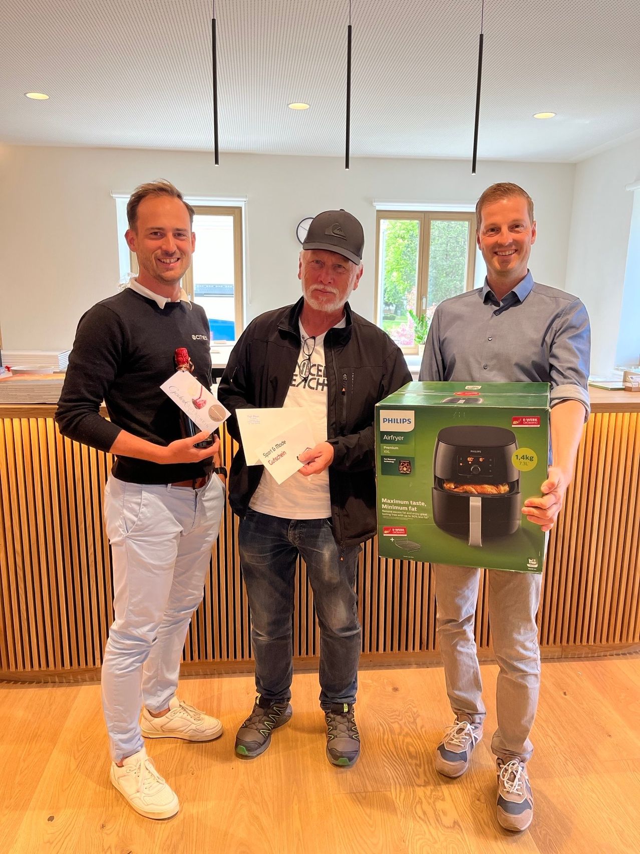 Drei Männer stehen drinnen zusammen. Einer hält eine Flasche, einer hält ein Papier und einer hält eine grüne Box mit einem schwarzen Gerät. Sie alle lächeln.
