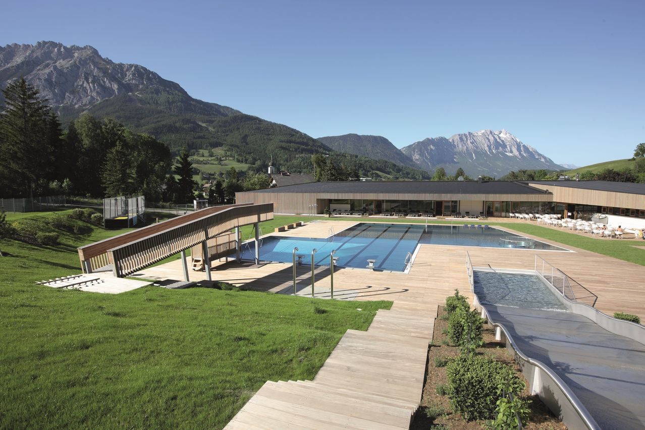 Luftaufnahme eines Freibadkomplexes mit Holzdeck und Bergen im Hintergrund.