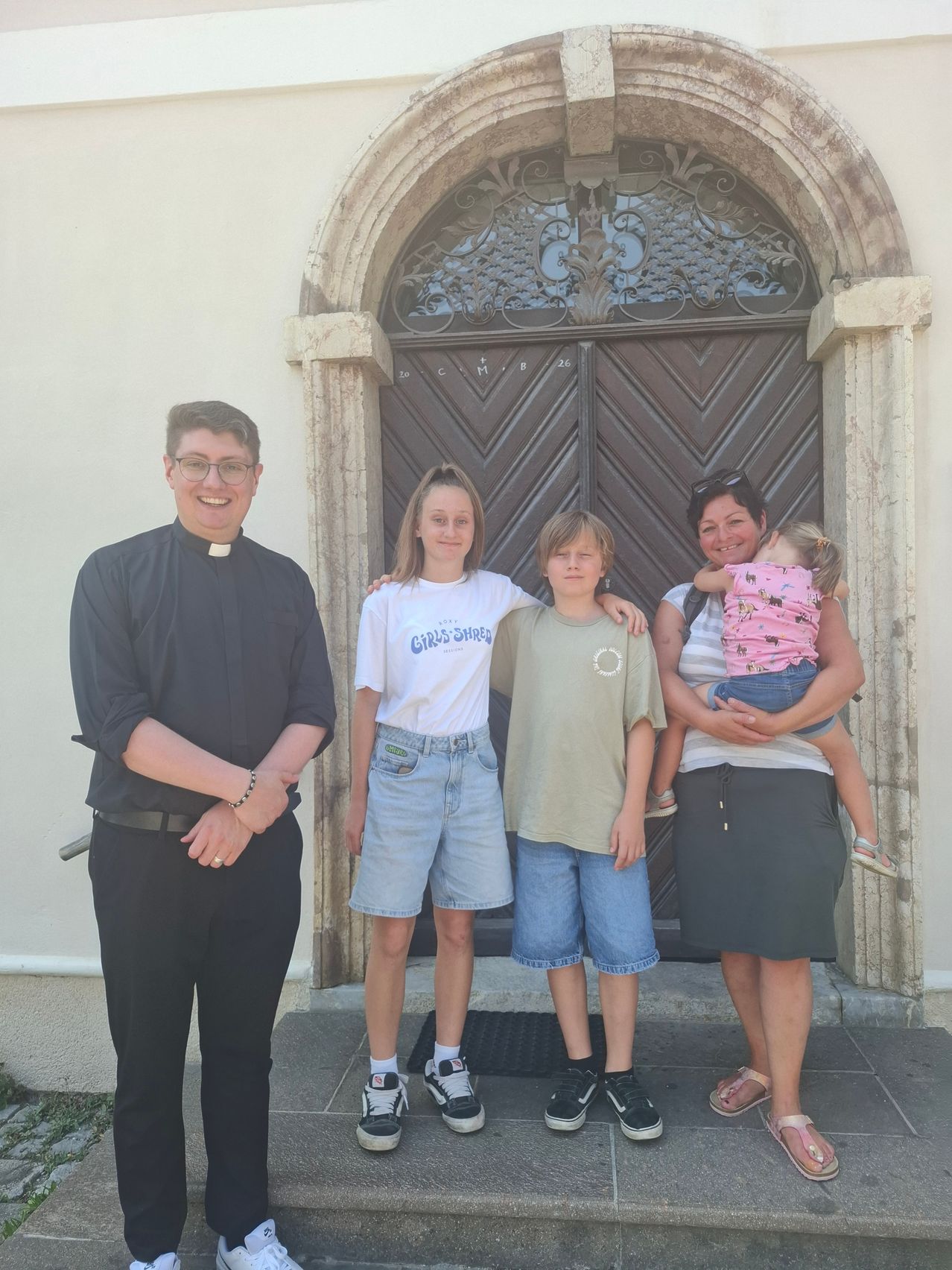 Ein Priester und eine Familie posieren vor einer Kirchentür. Der Mann trägt eine Brille und ein schwarzes Hemd. Die Frau hält ein Baby. Zwei Kinder stehen neben der Frau.