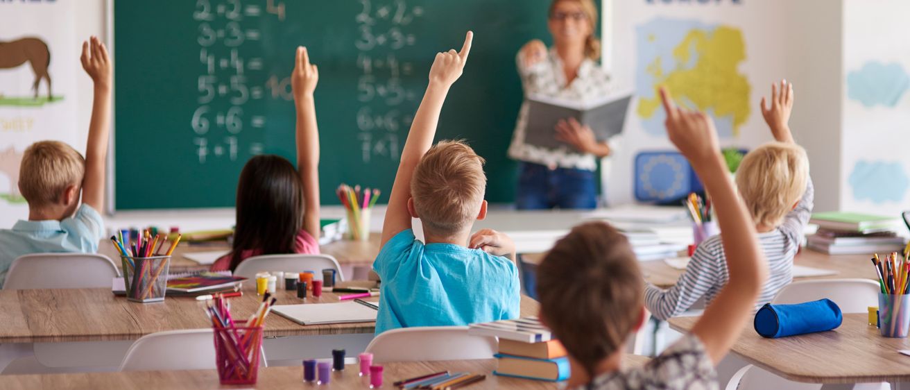 Ein Klassenzimmer mit Kindern, die ihre Hände heben, um eine Frage zu beantworten. Eine Lehrerin steht vor einer Tafel mit Matheproblemen.