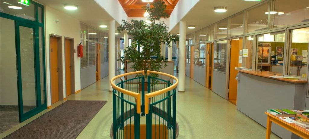 Ein Flur mit einer hölzernen Decke und einer grünen Brüstung um eine zentrale Pflanze, mit gläsernen Wänden auf beiden Seiten und einem Feuerlöscher an der Wand befestigt.