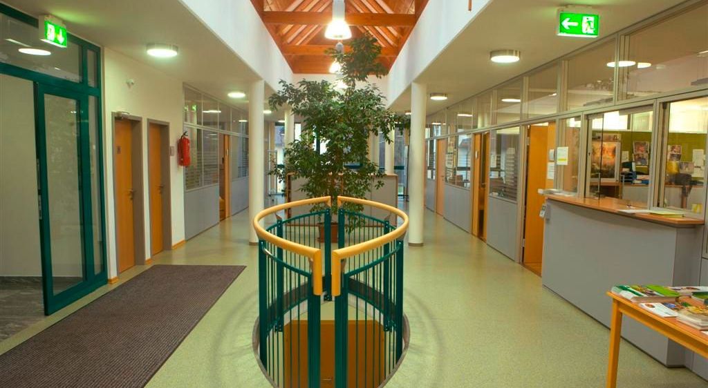 Ein Flur mit einer hölzernen Decke und einer grünen Brüstung um eine zentrale Pflanze, mit gläsernen Wänden auf beiden Seiten und einem Feuerlöscher an der Wand befestigt.