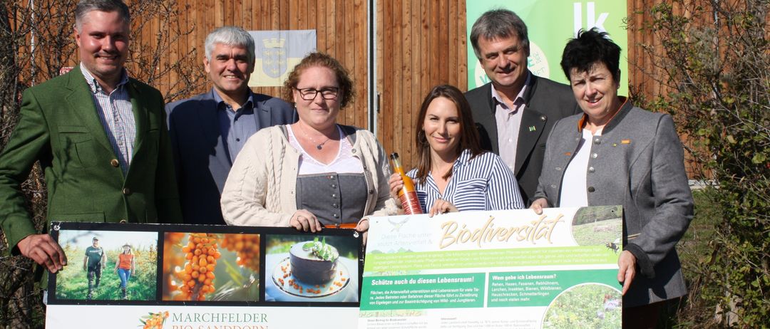 Eine Gruppe von Menschen steht draußen und posiert für ein Foto. Zwei Frauen halten ein Banner mit den Texten 'Biodiversitat' und 'Marchfelder Bio-Sanddorn'. Sie lächeln. Dahinter ist eine Holzwand und ein grünes Banner zu sehen.