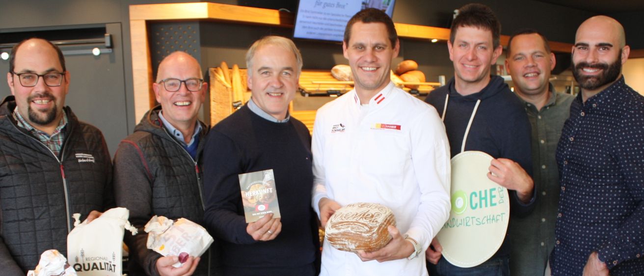 Sieben Männer posieren lächelnd zusammen und halten Lebensmittel in der Hand, einer von ihnen hält ein Schild mit der Aufschrift 'CHE'.