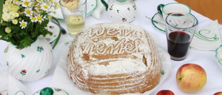 Ein Tisch mit Brot, Getränken und Obst. Das Brot trägt die Aufschrift 'Weine Weinel'. Zwei Äpfel, ein Glas Honig und eine Vase mit Blumen schmücken den Tisch.