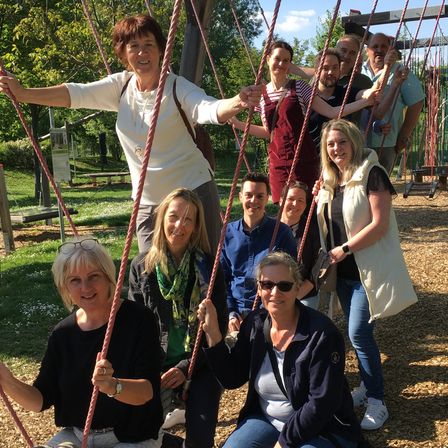Eine Gruppe Erwachsener posiert für ein Foto auf einer Schaukel in einem Park. Einige stehen, andere sitzen. Bäume und eine Holzbank sind im Hintergrund.