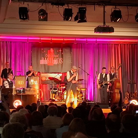 Eine Band spielt auf der Bühne, während ein Publikum zuschaut. Die Band hat ein Mikrofon und ein Banner hinter sich, auf dem Old School Bastards steht.
