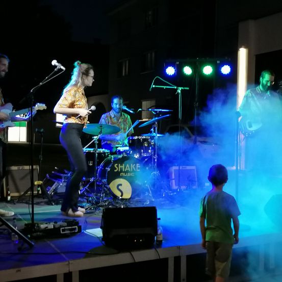 Eine Band tritt bei Nacht auf einer Bühne auf, während ein junger Junge zusieht. Die Bühne ist mit blauen und grünen Lichtern beleuchtet, und der Schlagzeuger hat ein großes Schlagzeugset.