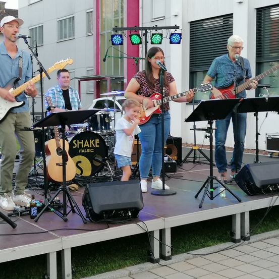 Eine Band mit einer Sängerin und Musikern tritt auf einer Bühne im Freien auf. Es gibt Lichter, Lautsprecher und ein Schlagzeug auf der Bühne.