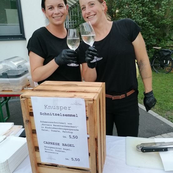 Zwei Frauen in Schwarz halten Weingläser draußen. Ein Holzkasten hat Schilder für 'Knusper Schnitzelsemmel' und 'Caprese Bagel'. Ein Tisch hat Teller, Besteck und ein Fahrrad.
