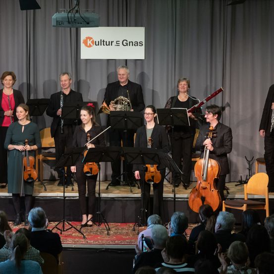 Ein Streichorchester spielt auf der Bühne mit Geigern, Cellisten und anderen Musikern, begleitet von einem Schild mit der Aufschrift 'Kultur & Gnas'. Das Publikum sitzt vor der Bühne und verfolgt die Aufführung.