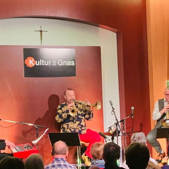 Zwei Musiker spielen Trompeten bei Kultur Gnas. Ein Publikum sitzt vor ihnen. An der Wand dahinter befindet sich ein Kreuz und ein Kultur Gnas-Schild.