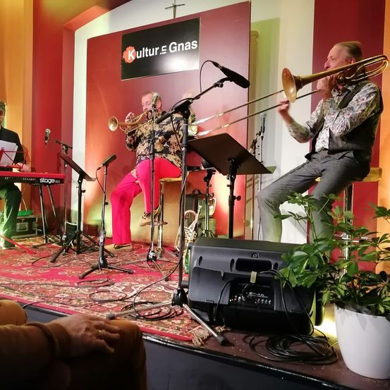 Drei Musiker spielen auf der Bühne. Einer spielt Posaune, ein anderer Trompete und der dritte Klavier. Die Bühne hat einen Teppich, ein Schild mit dem Text Kultur Gnäs und eine Pflanze.
